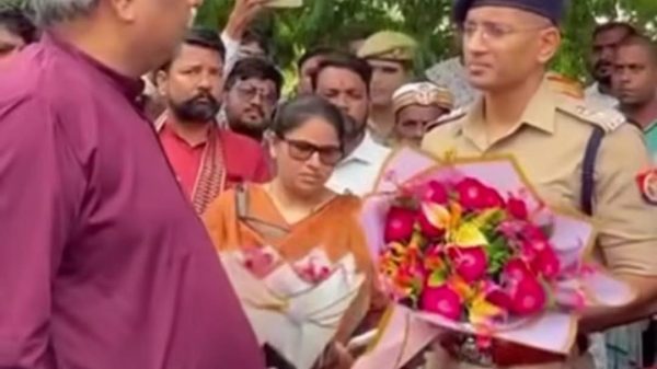 Man in a maroon kurta hands a bouquet to a uniformed police officer as a crowd looks on during an outdoor event.