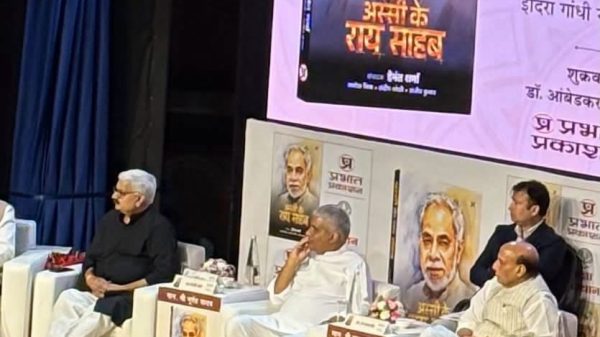 Panel of men seated on a stage during a book launch, with large posters of the author behind them.