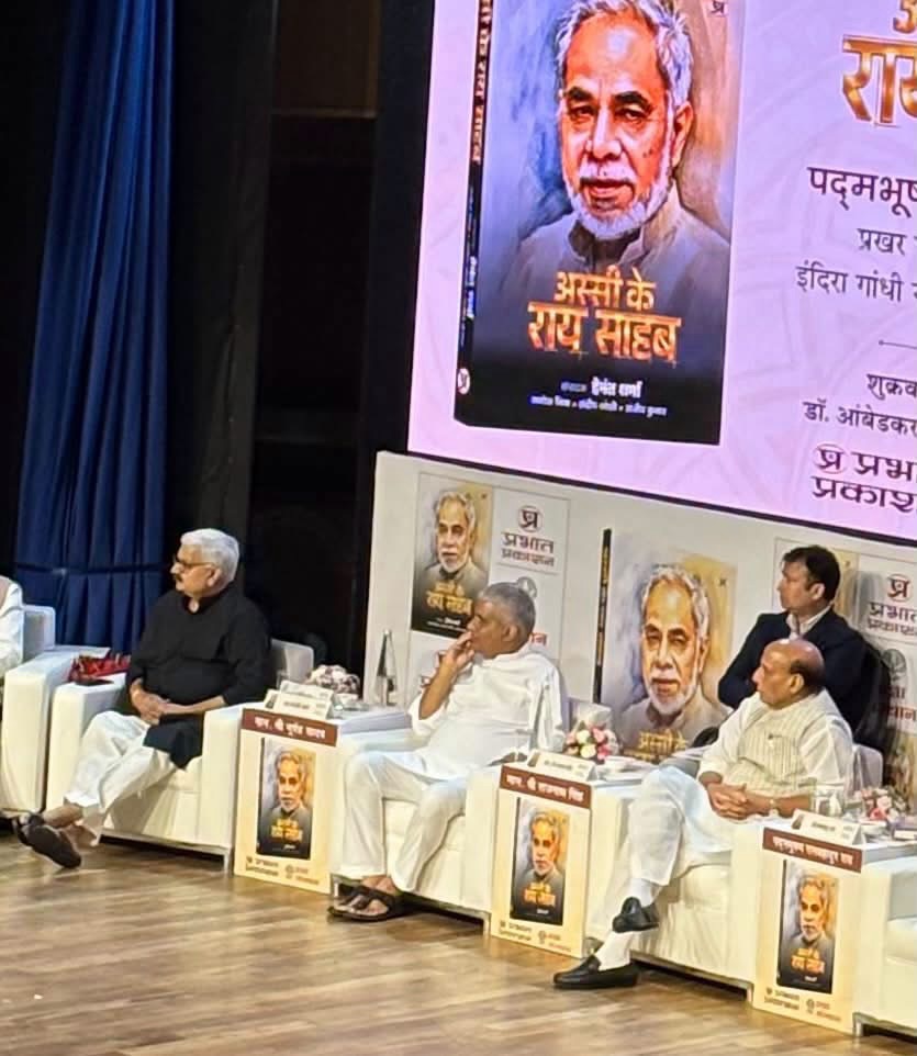 Panel of men seated on a stage during a book launch, with large posters of the author behind them.