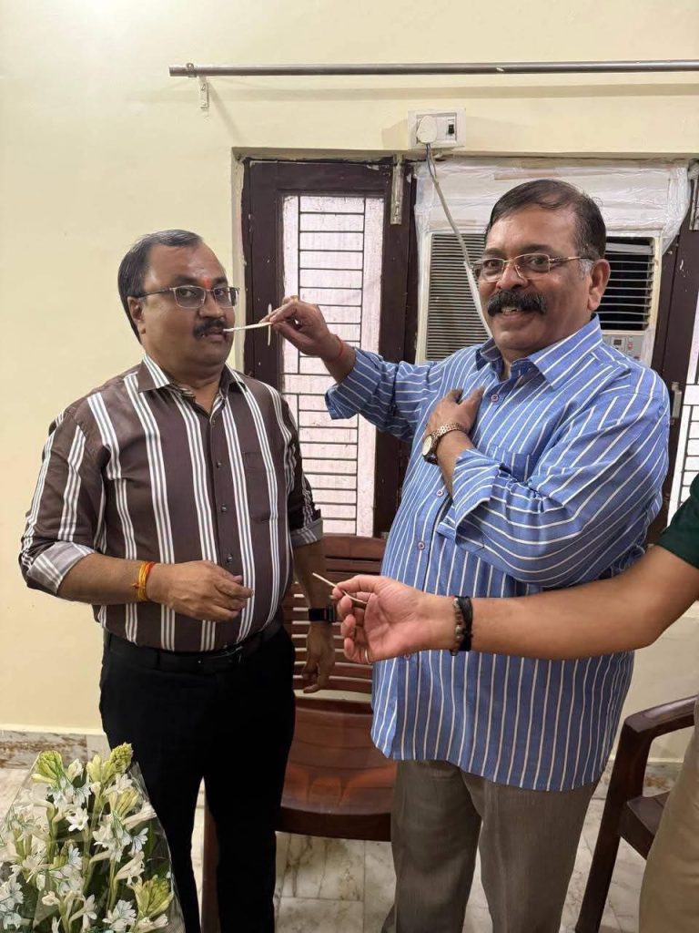 Two men indoors smiling as one holds a small stick near the other's mouth, with a bouquet visible in the foreground.