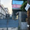 Blue metal gate with a yellow flower garland across it, closing a narrow alley beside a modern glass-front building with a pink sign reading 'NEWS' in the background.
