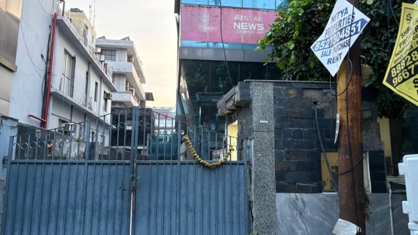 Blue metal gate with a yellow flower garland across it, closing a narrow alley beside a modern glass-front building with a pink sign reading 'NEWS' in the background.