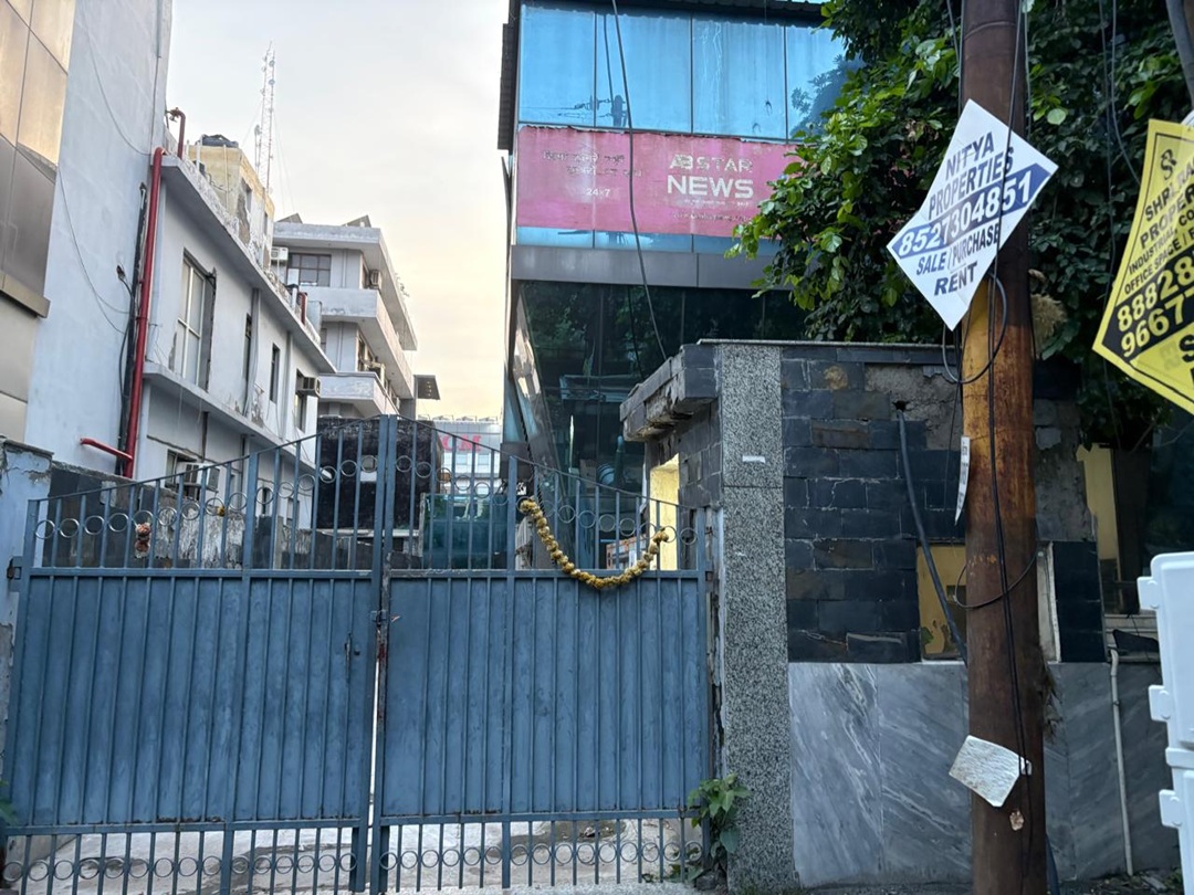 Blue metal gate with a yellow flower garland across it, closing a narrow alley beside a modern glass-front building with a pink sign reading 'NEWS' in the background.