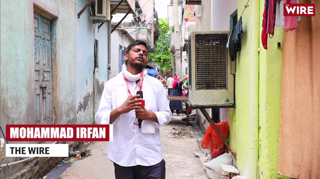 Mohammad Irfan, a The Wire reporter, stands in a narrow alley holding a microphone in a white shirt.