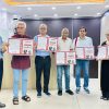 Six adults in an office pose together, each holding an open red-framed booklet with text and images on the pages.