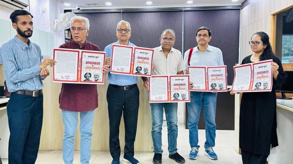 Six adults in an office pose together, each holding an open red-framed booklet with text and images on the pages.
