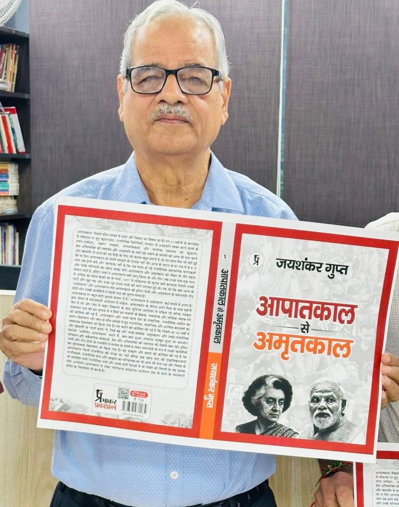 Older man with glasses holding up a large, red-bordered booklet featuring Hindi text in an office setting, facing the camera.