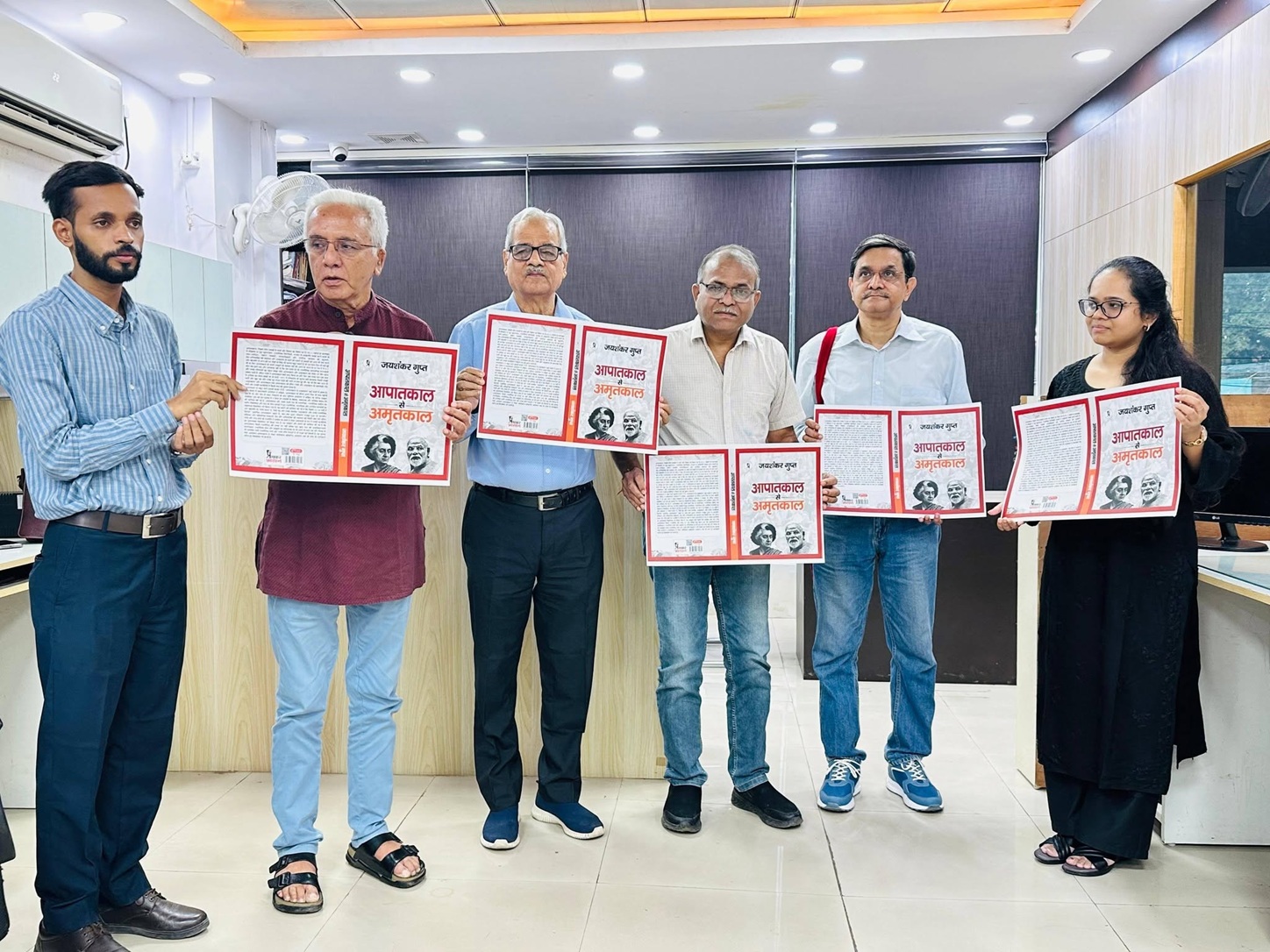 Six adults in an office pose together, each holding an open red-framed booklet with text and images on the pages.