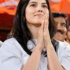 Young woman in a white shirt with hands pressed together in a prayer-like gesture, smiling at an event.