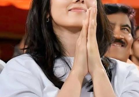 Young woman in a white shirt with hands pressed together in a prayer-like gesture, smiling at an event.