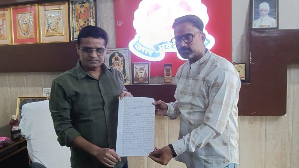 lahiri tk_1 | No. 1 Indian Media News Portal Two men stand in an office exchanging a document, with framed awards and a red sign behind them, creating a formal scene.