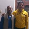 Two men standing indoors, one with his arm around the other; left man wears a blue sweater and beige vest, right man in a yellow hoodie.