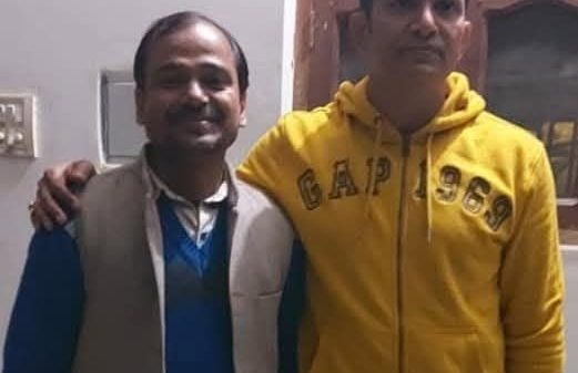Two men standing indoors, one with his arm around the other; left man wears a blue sweater and beige vest, right man in a yellow hoodie.