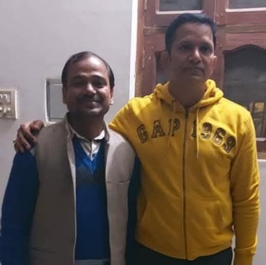 Two men standing indoors, one with his arm around the other; left man wears a blue sweater and beige vest, right man in a yellow hoodie.