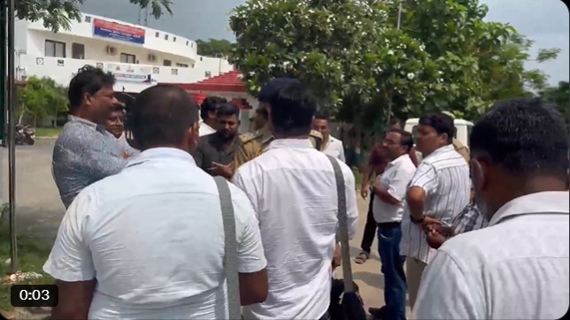 A group of men in white shirts stand in a circle outdoors, talking near a campus or government building with trees in the background (video still).