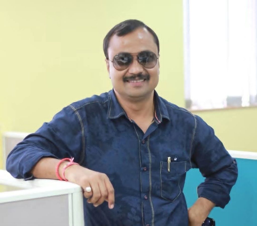 Man in a denim shirt and sunglasses leaning on a desk in a bright office, smiling at the camera.