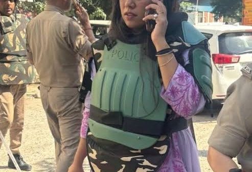 Woman in a light purple outfit wearing green police body armor, talking on a cellphone and holding a helmet, with uniformed officers and a dusty street in the background.
