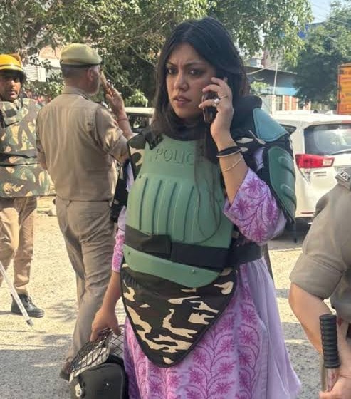 Woman in a light purple outfit wearing green police body armor, talking on a cellphone and holding a helmet, with uniformed officers and a dusty street in the background.