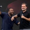 Two men on a stage share a hug while speaking into microphones; one points to the audience, both smiling.