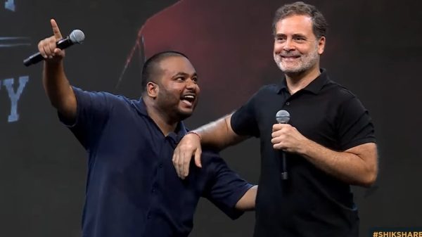 Two men on a stage share a hug while speaking into microphones; one points to the audience, both smiling.