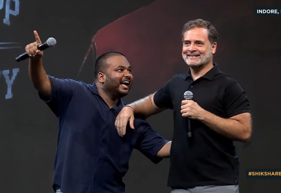 Two men on a stage share a hug while speaking into microphones; one points to the audience, both smiling.
