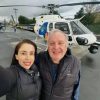 Two people in jackets posing for a selfie on a tarmac with a white helicopter in the background, smiling at the camera