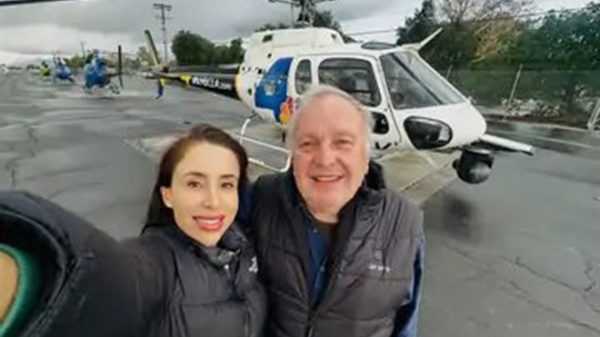 Two people in jackets posing for a selfie on a tarmac with a white helicopter in the background, smiling at the camera