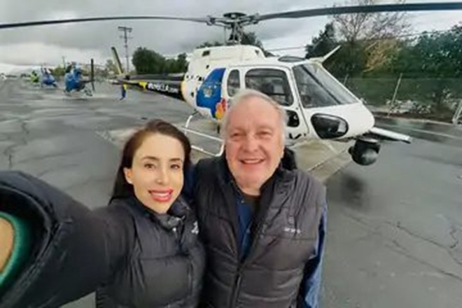 Two people in jackets posing for a selfie on a tarmac with a white helicopter in the background, smiling at the camera
