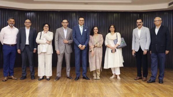 Nine professionals in business attire pose for a group photo in a room with a dark wood-paneled wall, on a wooden floor.
