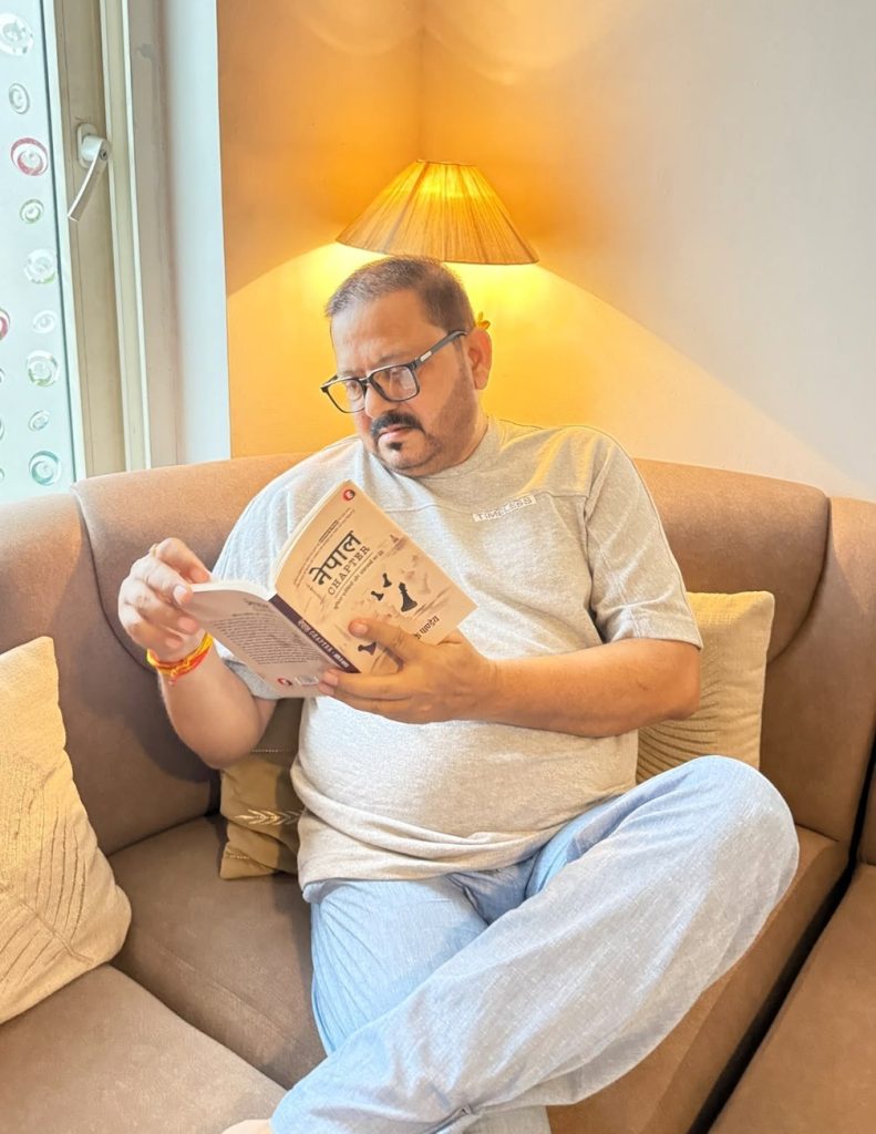 Man with glasses sits on a beige sofa, reading a paperback by a warm lamp.