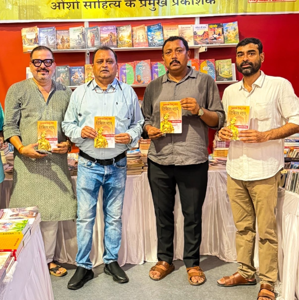 Four men stand in a bookstore display, each holding a yellow-covered book with a man on the cover, shelves of books behind them.