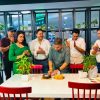 A man cuts a cake at a table while colleagues clap and smile in a bright office space with plants in the background.