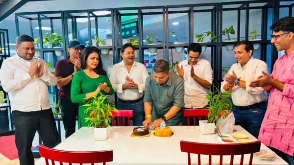 A man cuts a cake at a table while colleagues clap and smile in a bright office space with plants in the background.
