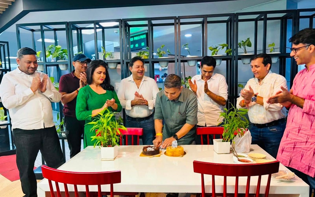 A man cuts a cake at a table while colleagues clap and smile in a bright office space with plants in the background.