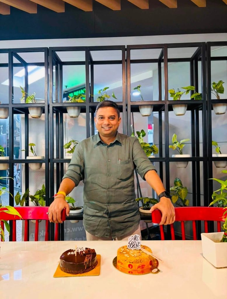 nripendra singh | No. 1 Indian Media News Portal Man in a green shirt stands behind a white table with two cakes in front, indoors with a plant-filled black grid backdrop behind him.