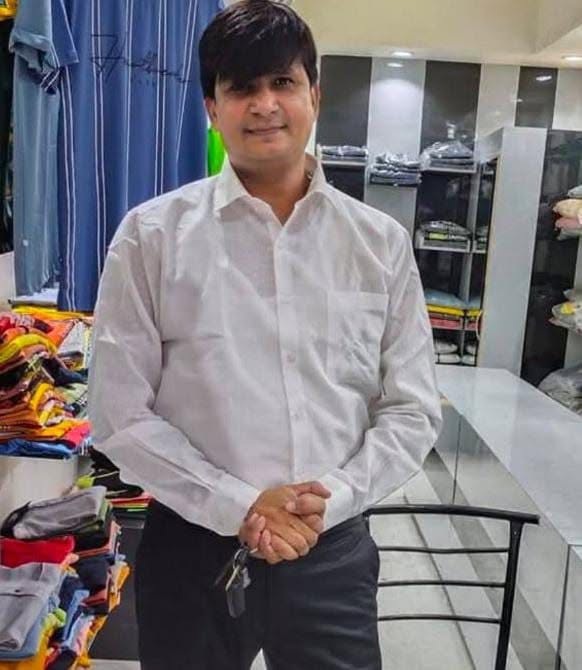 om aditya | No. 1 Indian Media News Portal Man in a white dress shirt and dark pants standing in a clothing store, folding his hands with folded garments in the background