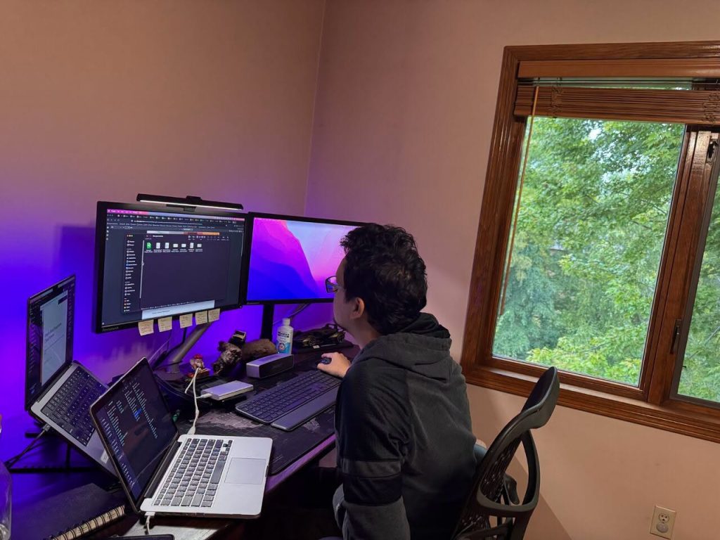 Person in a hoodie sits at a desk with multiple monitors and a laptop, coding in a home office with purple ambient lighting on the walls.