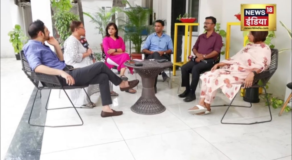 Six people sitting in a circle on a patio, appearing to be a panel discussion for News18 India, with plants in the background and a coffee table in the center.