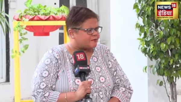 Woman wearing glasses and a patterned blouse is being interviewed, speaking into a News18 microphone outdoors with a potted plant in the background.
