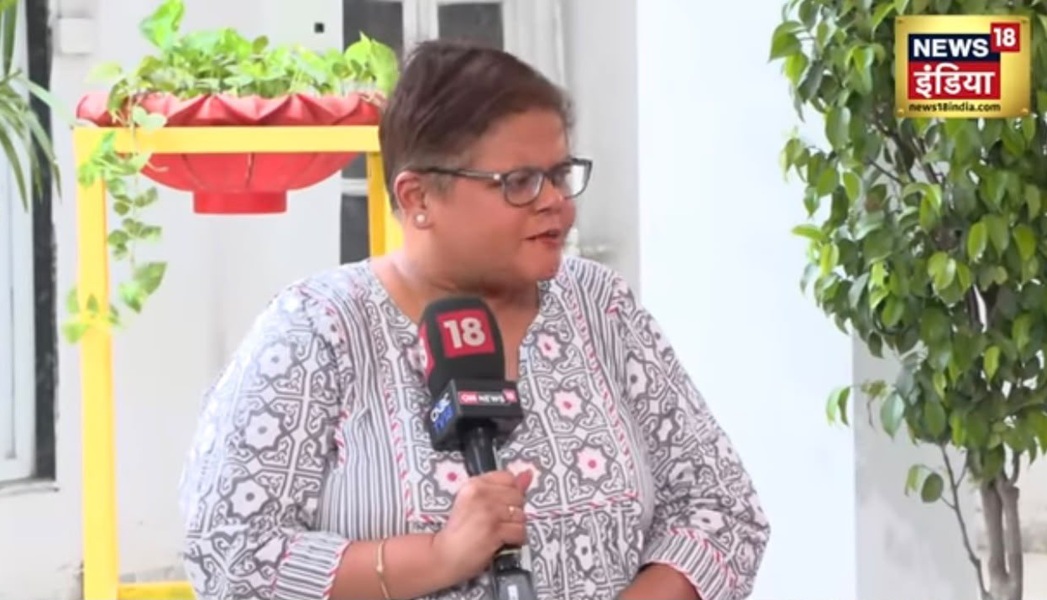 Woman wearing glasses and a patterned blouse is being interviewed, speaking into a News18 microphone outdoors with a potted plant in the background.