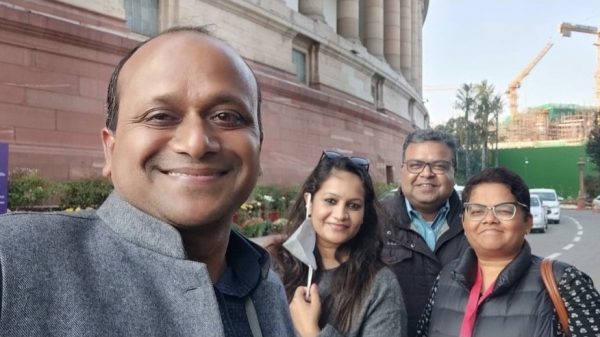 Four friends take a group selfie outside a large red-stone building, smiling at the camera.