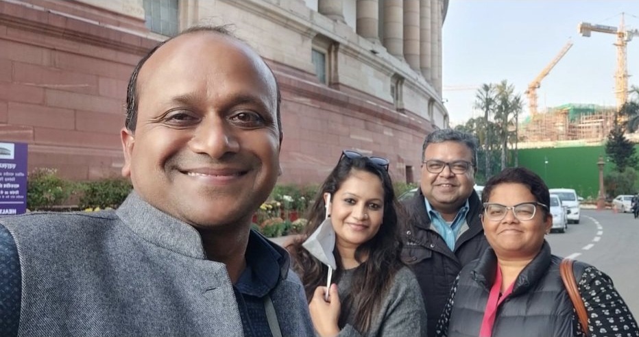 Four friends take a group selfie outside a large red-stone building, smiling at the camera.