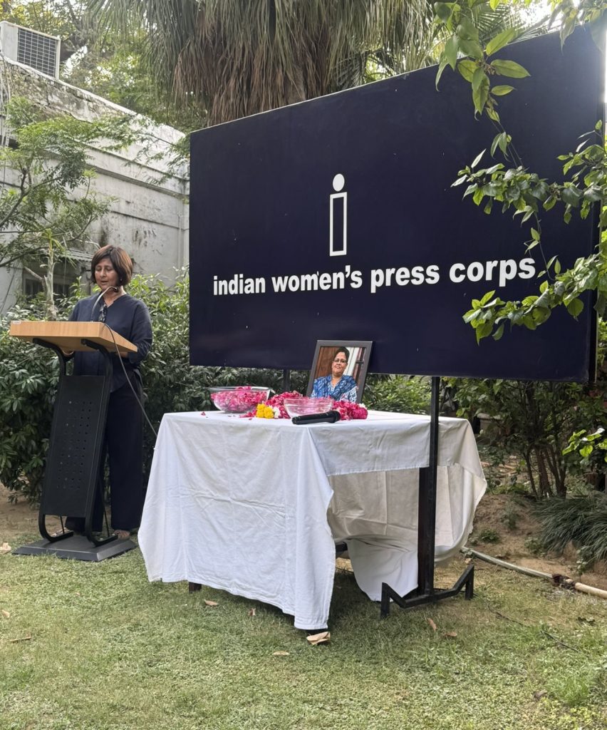 Woman speaks at a podium beside a large sign that reads 'indian women's press corps' at an outdoor memorial with a framed portrait and bowls of flowers on a table nearby.