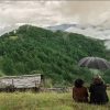 Two people sit on a grassy hilltop with a black umbrella, watching a misty green valley of pine forests and distant mountains.