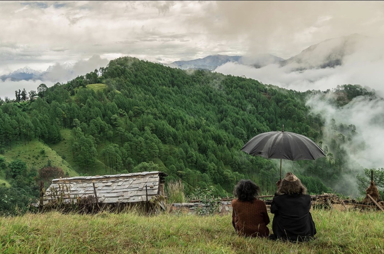 Two people sit on a grassy hilltop with a black umbrella, watching a misty green valley of pine forests and distant mountains.