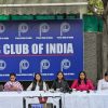 Woman standing and speaking into a microphone at a Press Club of India panel, with five women seated at a long table and a blue backdrop reading 'Press Club of India'.