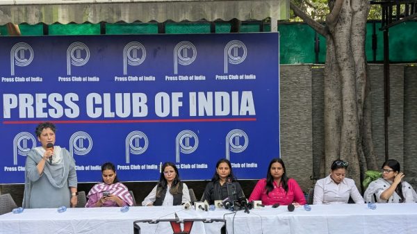 Woman standing and speaking into a microphone at a Press Club of India panel, with five women seated at a long table and a blue backdrop reading 'Press Club of India'.