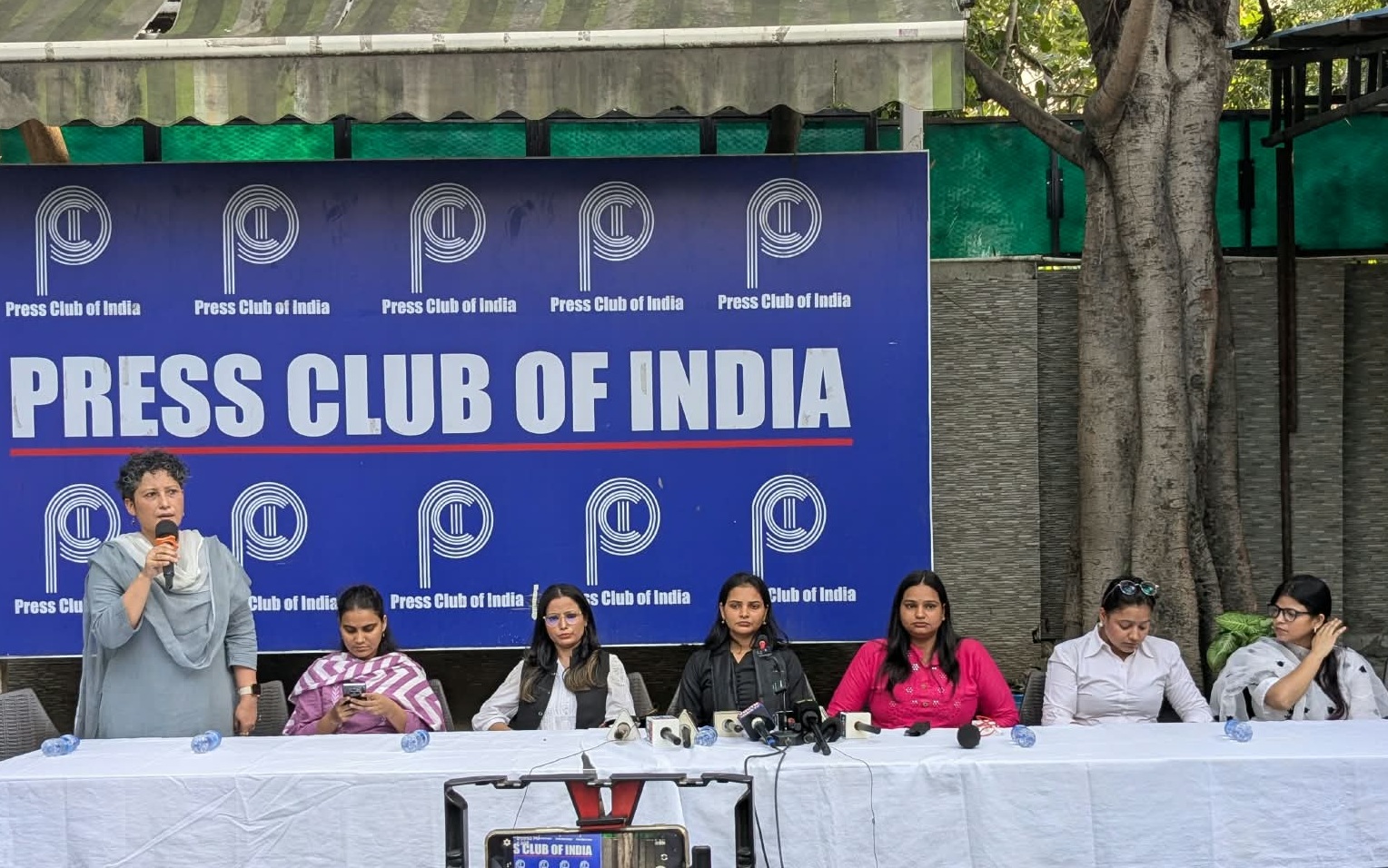 Woman standing and speaking into a microphone at a Press Club of India panel, with five women seated at a long table and a blue backdrop reading 'Press Club of India'.