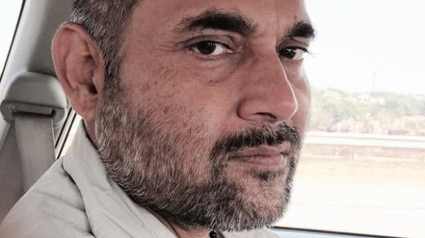 Man with stubble and short hair seated in a car, looking at the camera with a neutral expression.
