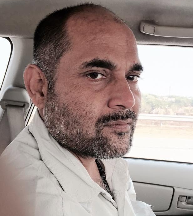 Man with stubble and short hair seated in a car, looking at the camera with a neutral expression.